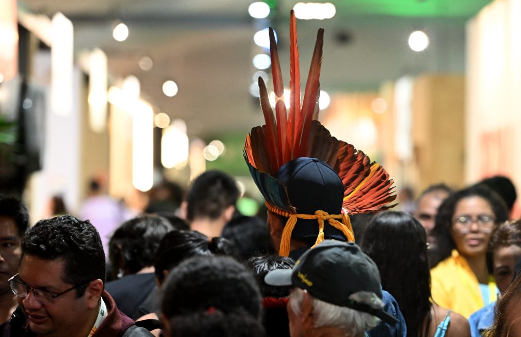 COP30 participants walking down a brightly-lit hallway, one person wearing a traditional Amazonian feathered headdress.