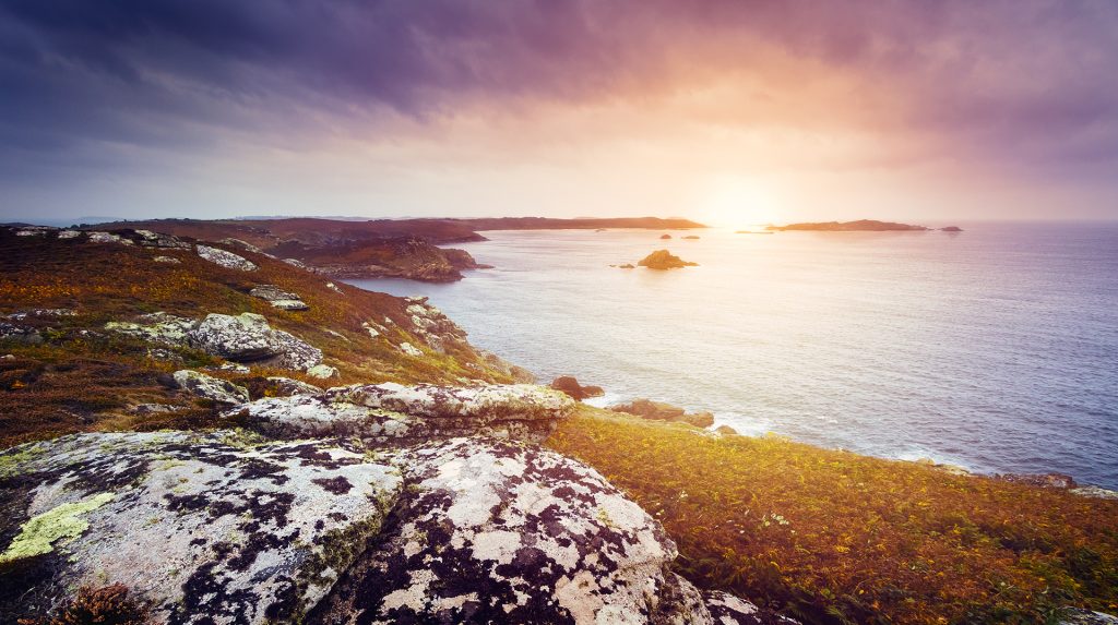 The sun rising over the ocean, with a rocky shoreline in the foreground.